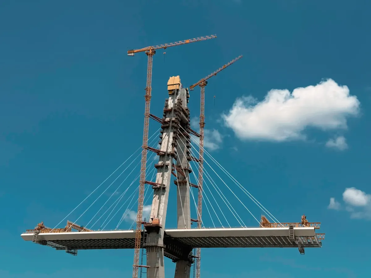 Dynamic view of a cable-stayed bridge under construction with cranes against a clear blue sky.