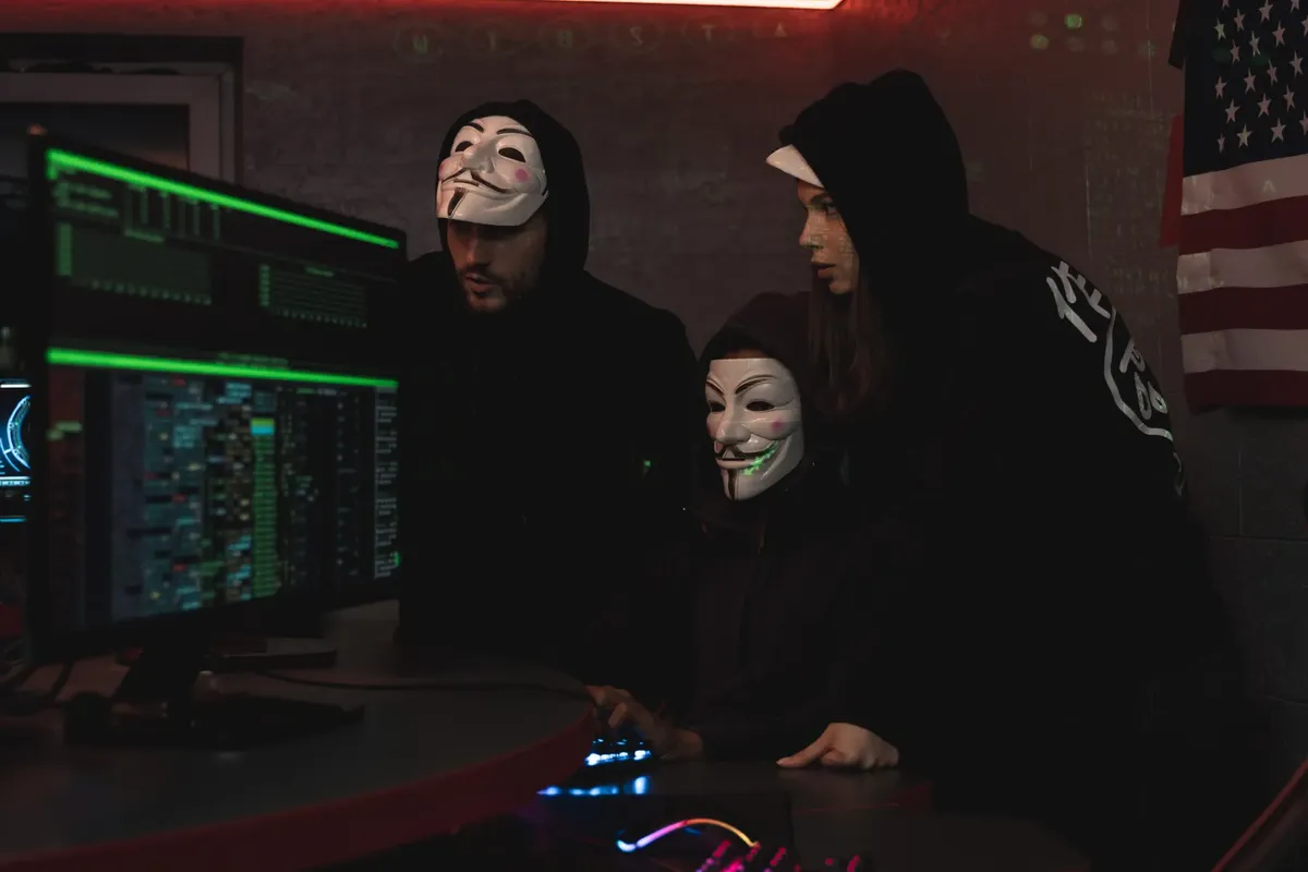 Masked hackers in hoodies working on a system in a dimly lit room, representing cyber security threats.